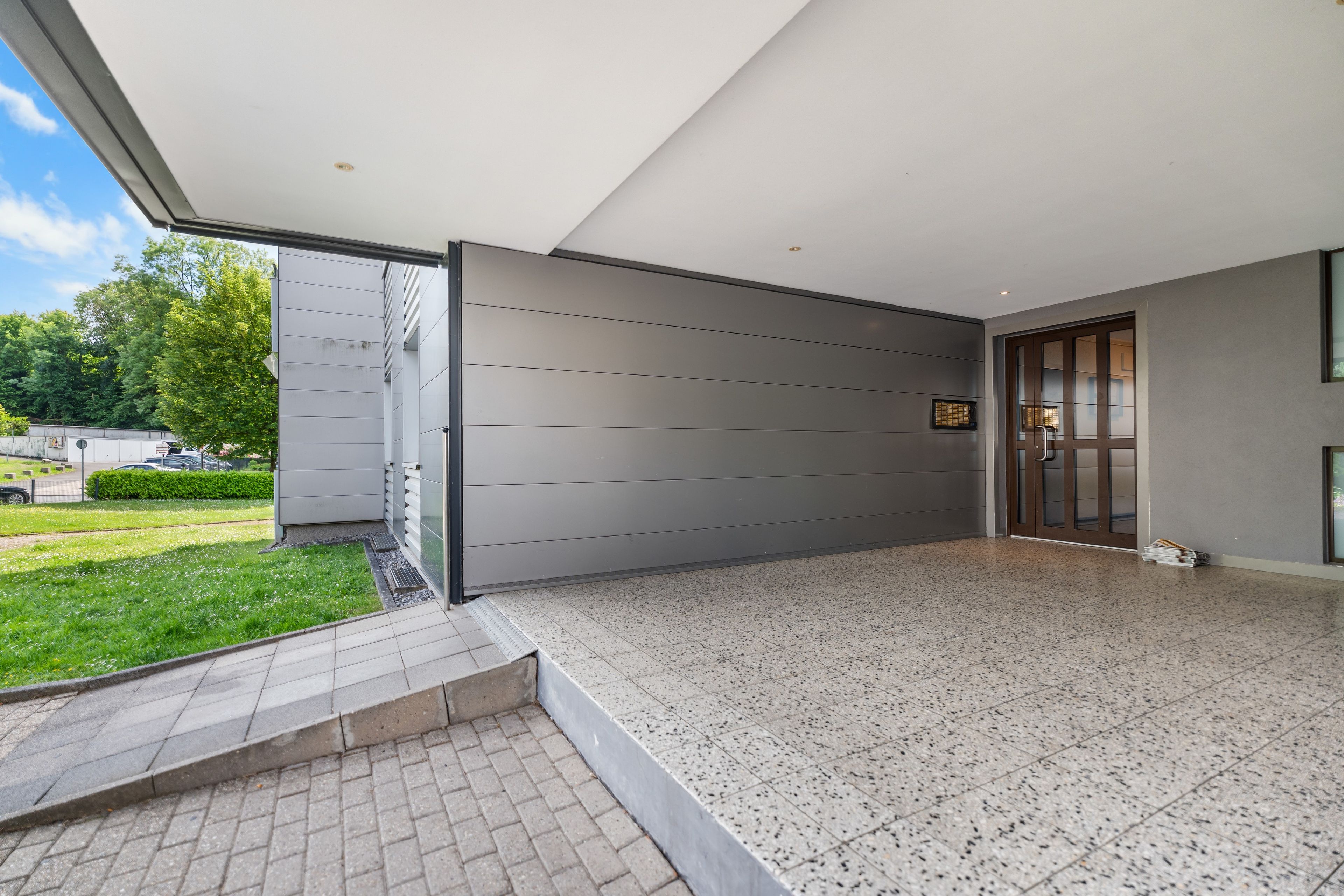 Un edificio moderno con un patio cubierto, entrada por puerta de vidrio y espacio exterior pavimentado.