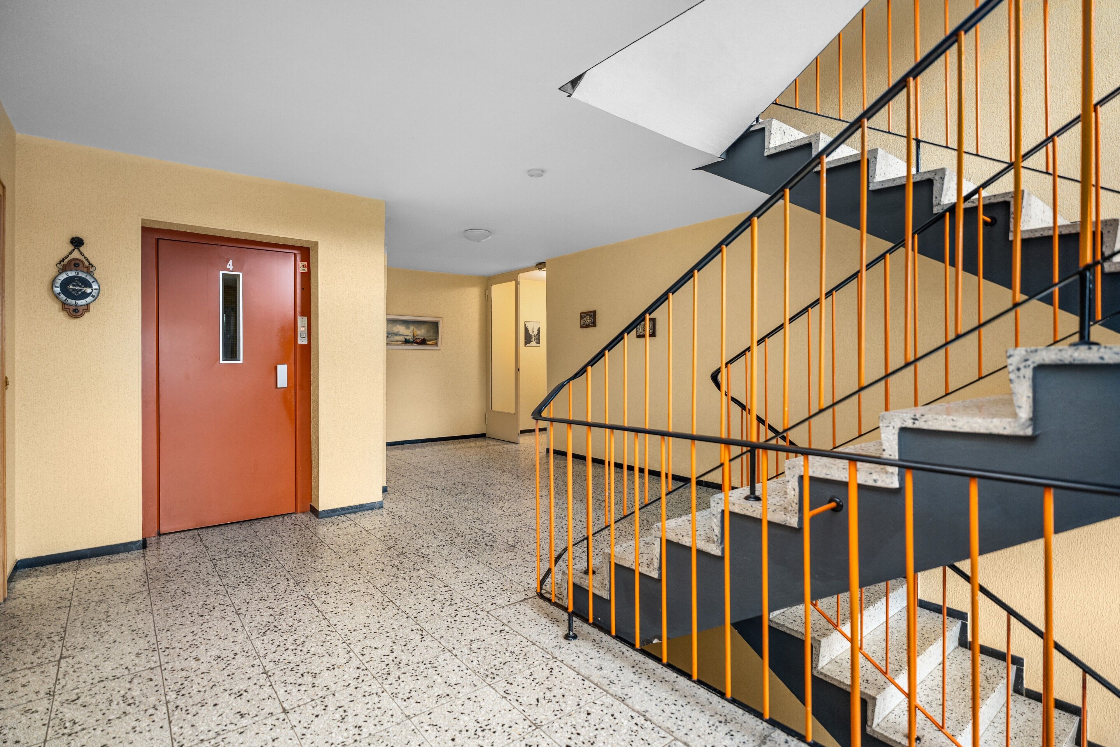 Ein Flur mit einer Tür, die mit '4' beschriftet ist, einer Treppe mit orangefarbenen Geländern und einem punktierten Boden.