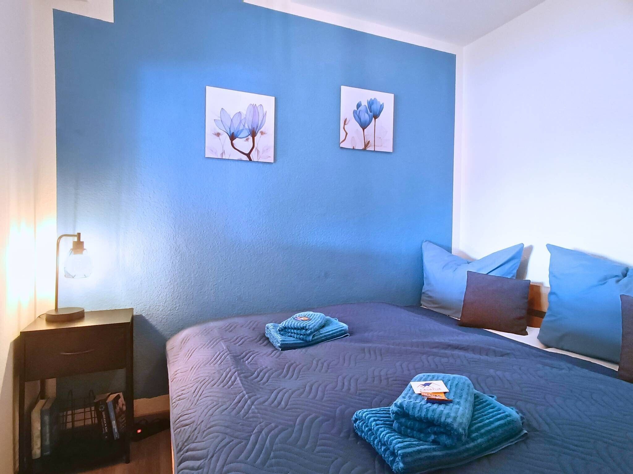 Ein Schlafzimmer mit einem großen Bett, zwei Kissen, einem Nachttisch mit einer Lampe und zwei an der Wand befestigten Bildern.