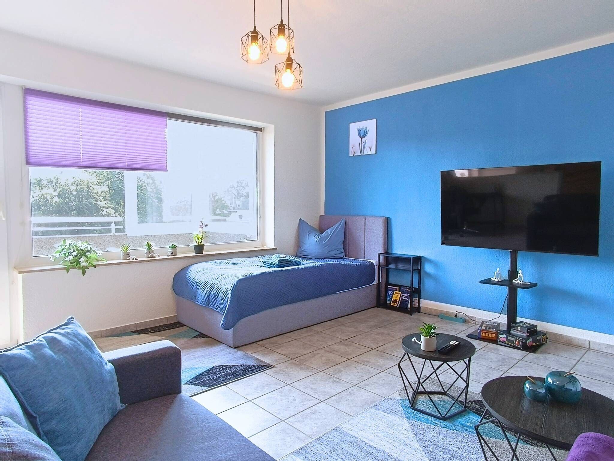 Une chambre moderne avec un lit, un canapé, une télévision et une fenêtre équipée de stores.