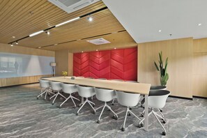 Een congrescentrum met een lange tafel, witte stoelen, een rode accentmuur en een projectiescherm.