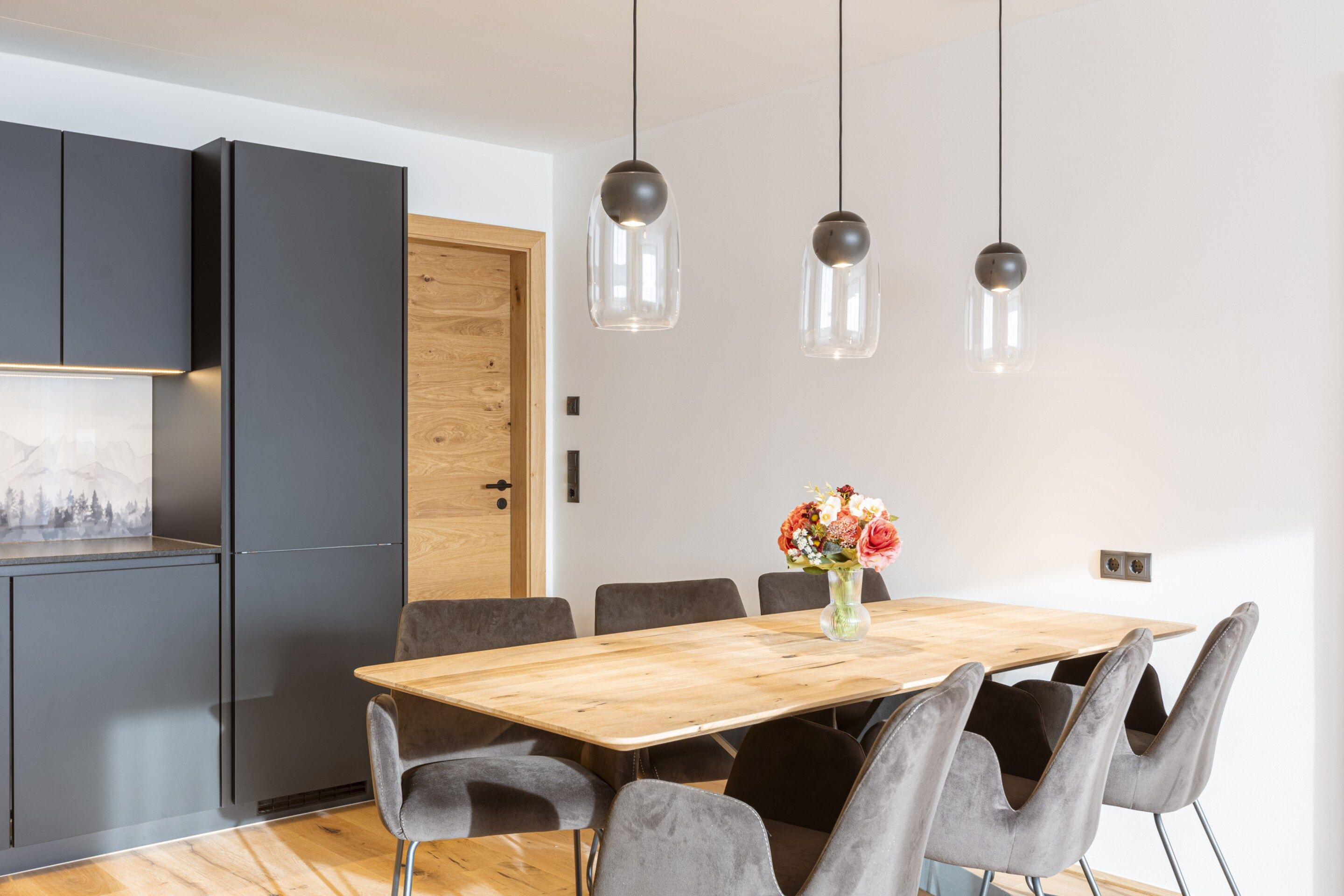 Un espace de salle à manger moderne avec une table en bois, des chaises à revêtement gris et des luminaires suspendus.