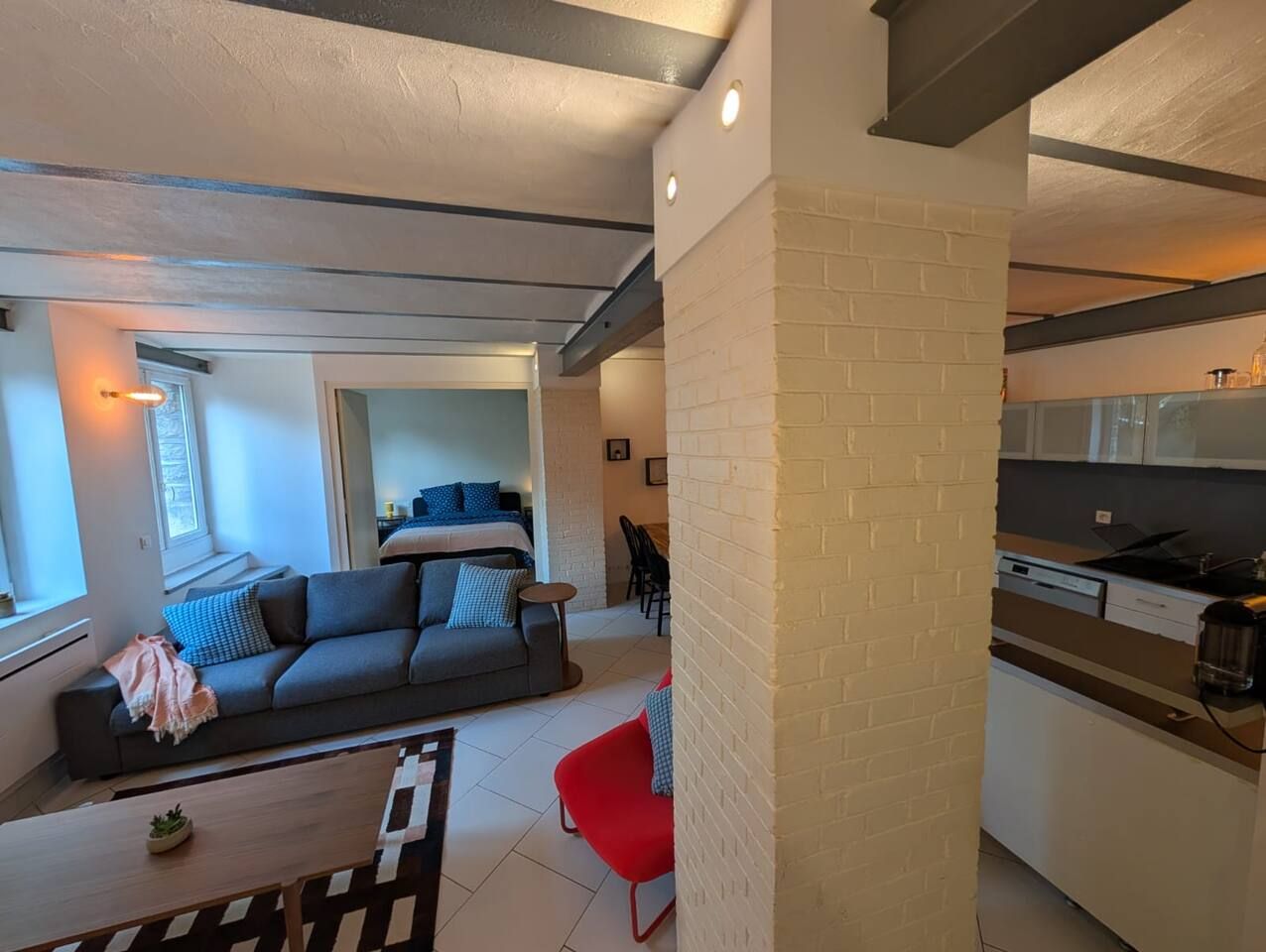 Ein modernes Wohnzimmer mit einer Couch, einem roten Sessel und einem Essbereich, wobei im Hintergrund eine Küche zu sehen ist.