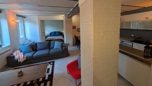 Ein modernes Wohnzimmer mit einer Couch, einem roten Sessel und einem Essbereich, wobei im Hintergrund eine Küche zu sehen ist.