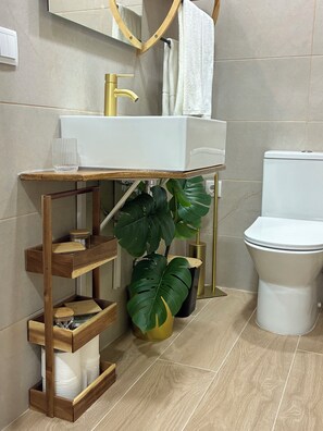 A bathroom with a white sink, gold faucet, wooden shelves, a potted plant, and a toilet. -  “Uma estadia com charme no coração de Torres Vedras”
Apartamento aconchegante (Torres Vedras)