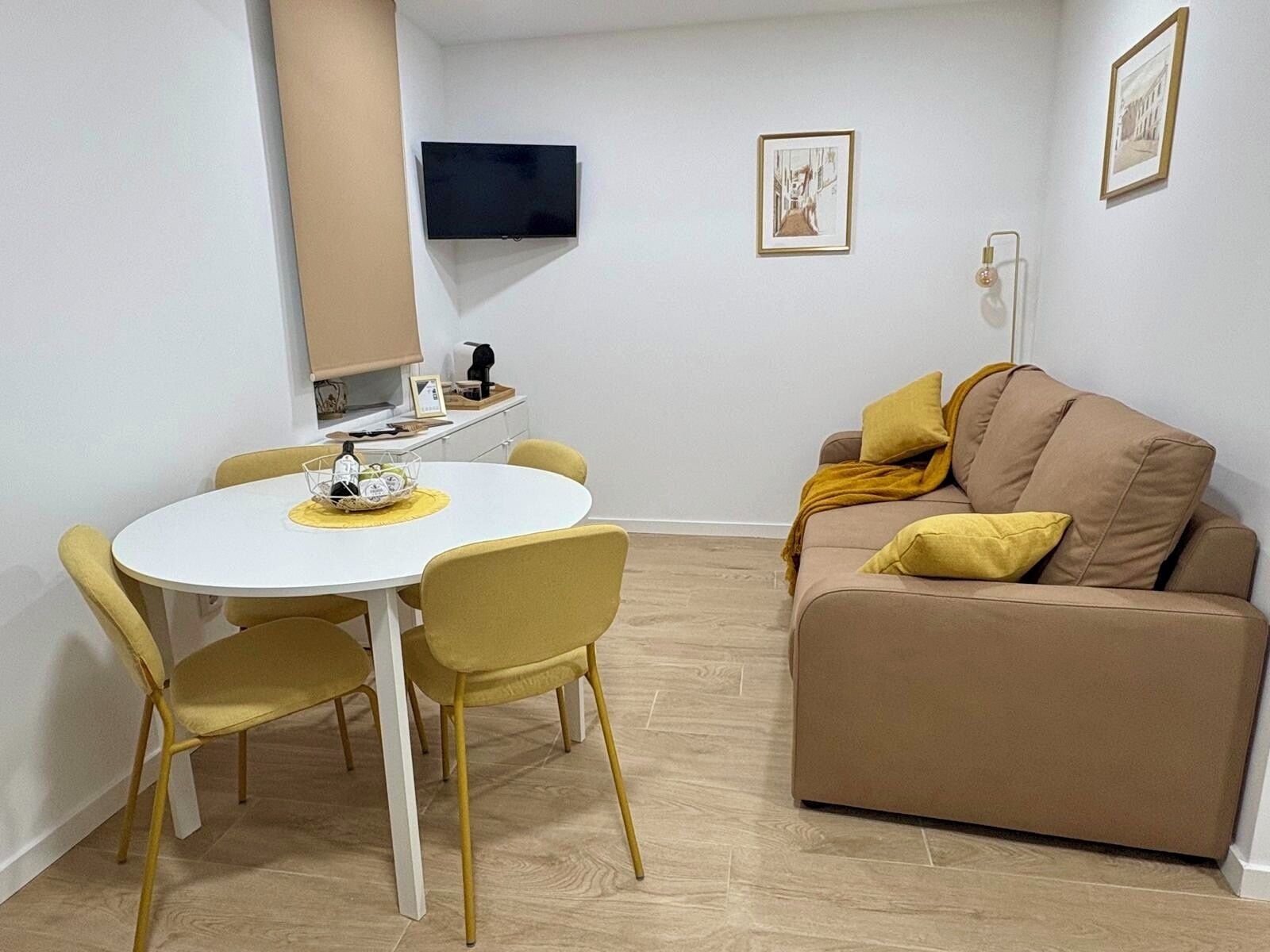 Una sala de estar moderna con una mesa redonda blanca, sillas amarillas, un sofá marrón con cojines amarillos, un televisor de pantalla plana montado en la pared y un cuadro enmarcado.