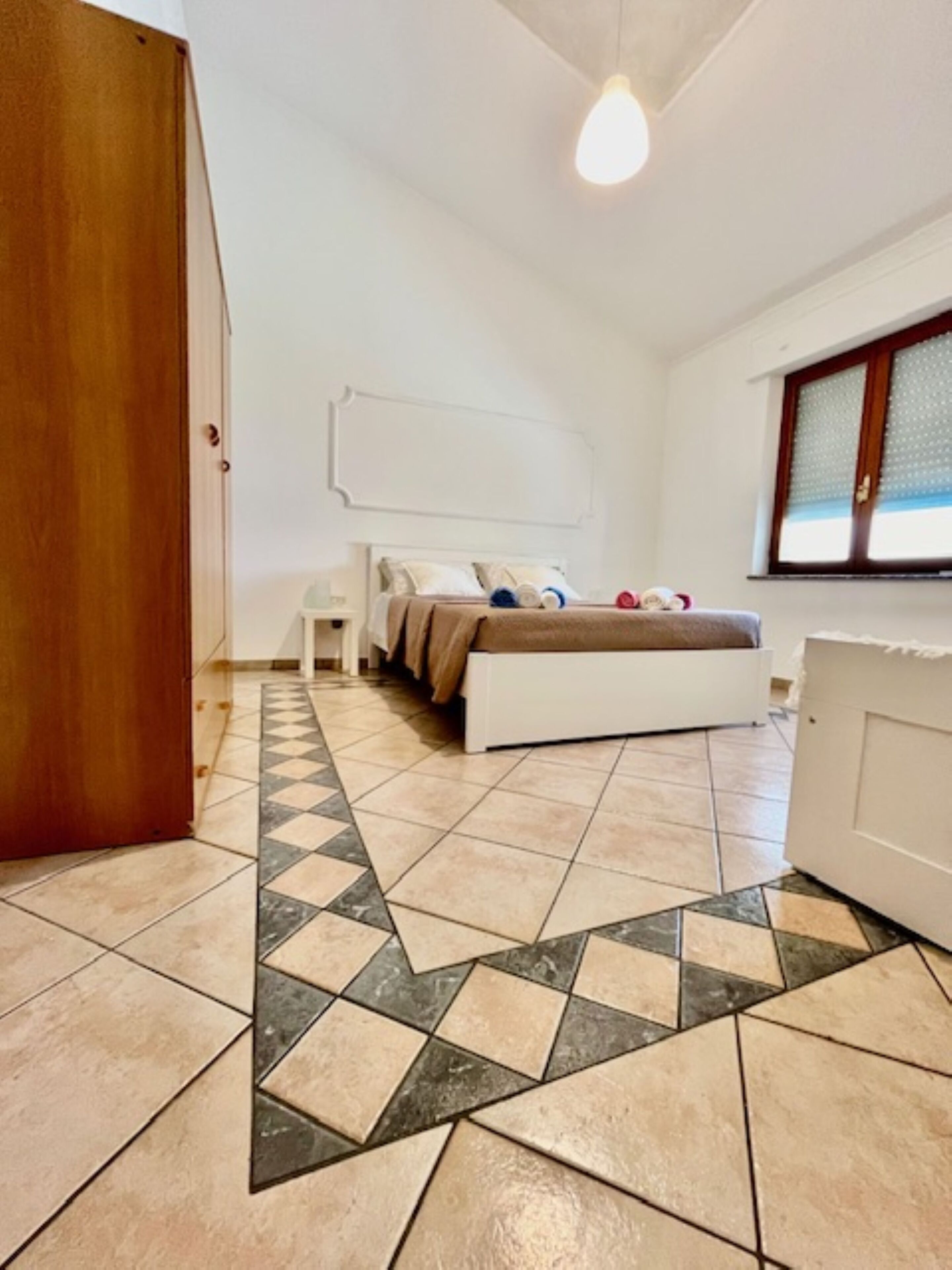 Een slaapkamer met een bed, nachtkastje, dressoir en raam met jaloezieën.