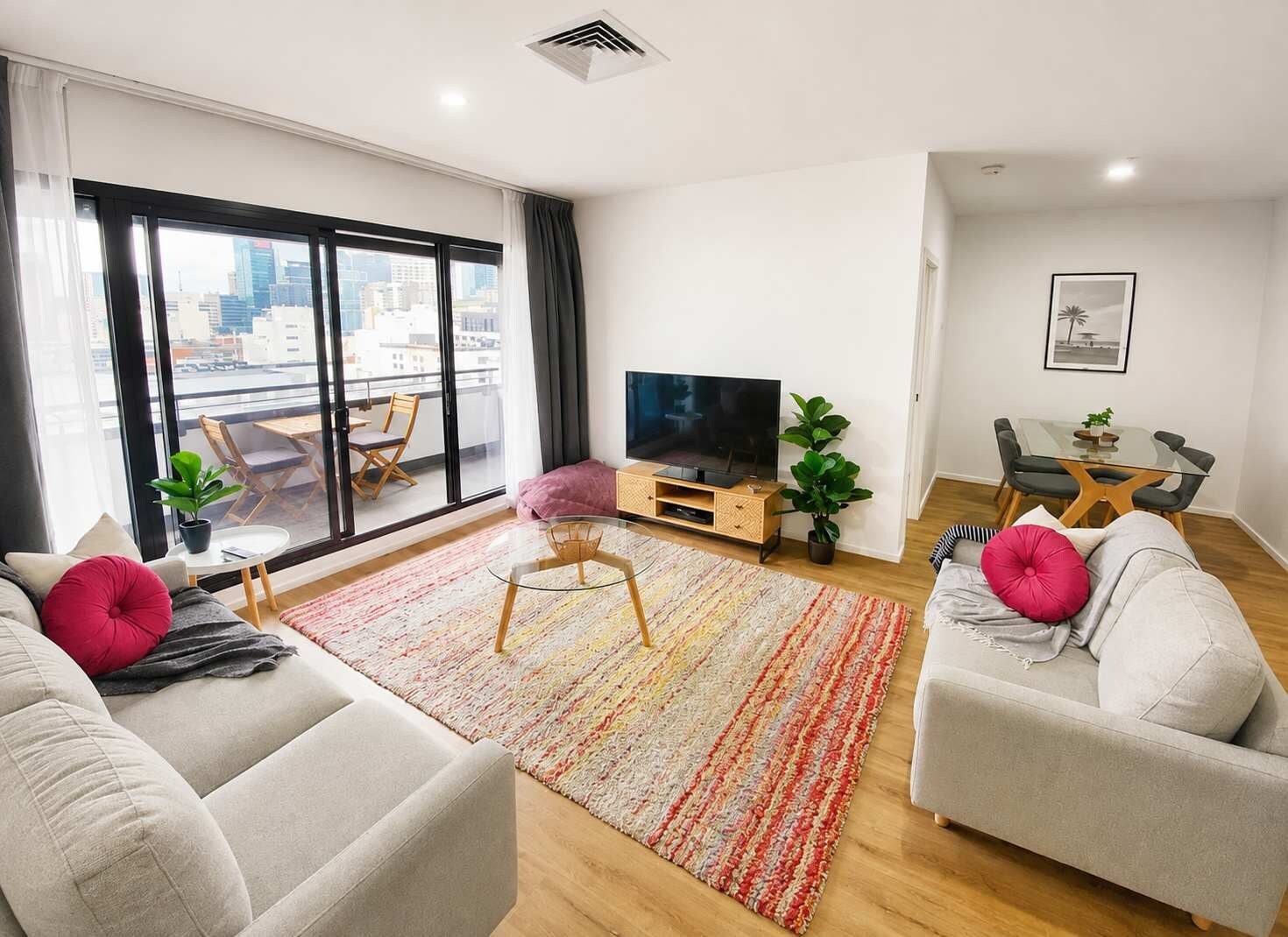 A modern living room with a large window, a flat-screen TV, a dining area, and two sofas.