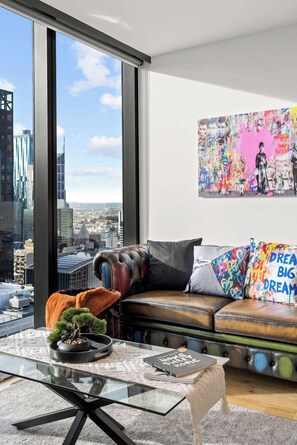 Een moderne woonkamer met uitzicht over de stad, een glazen salontafel, een leren bank en een kleurrijk abstract schilderij.