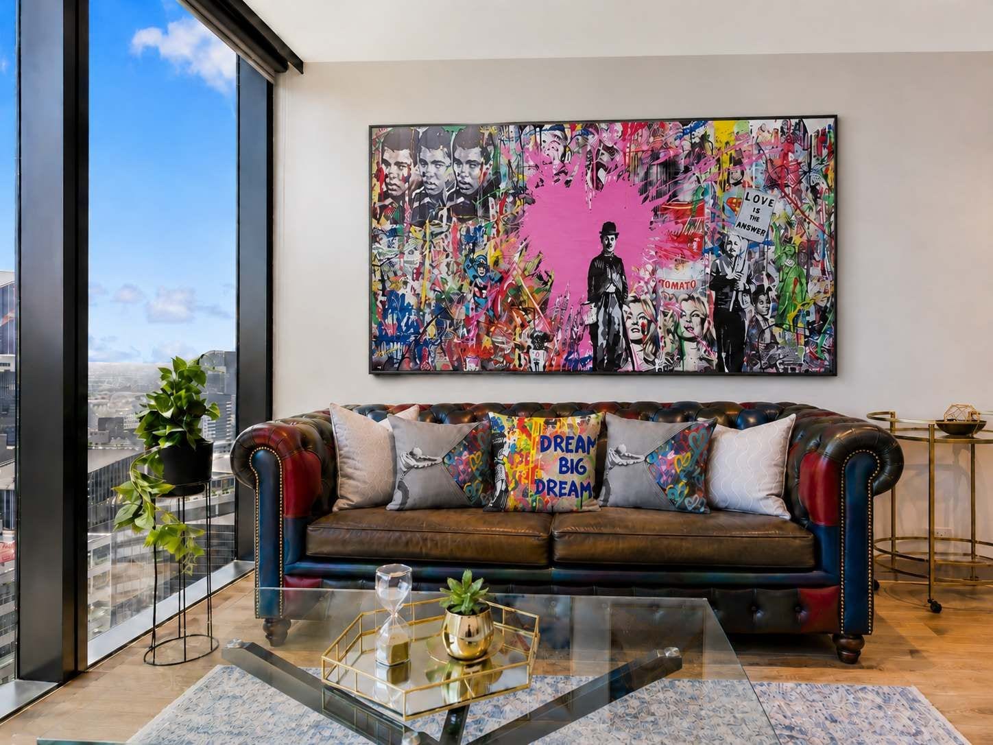 Un salon moderne avec un canapé en cuir, des coussins colorés, une table basse en verre et un grand tableau abstrait accroché au mur.