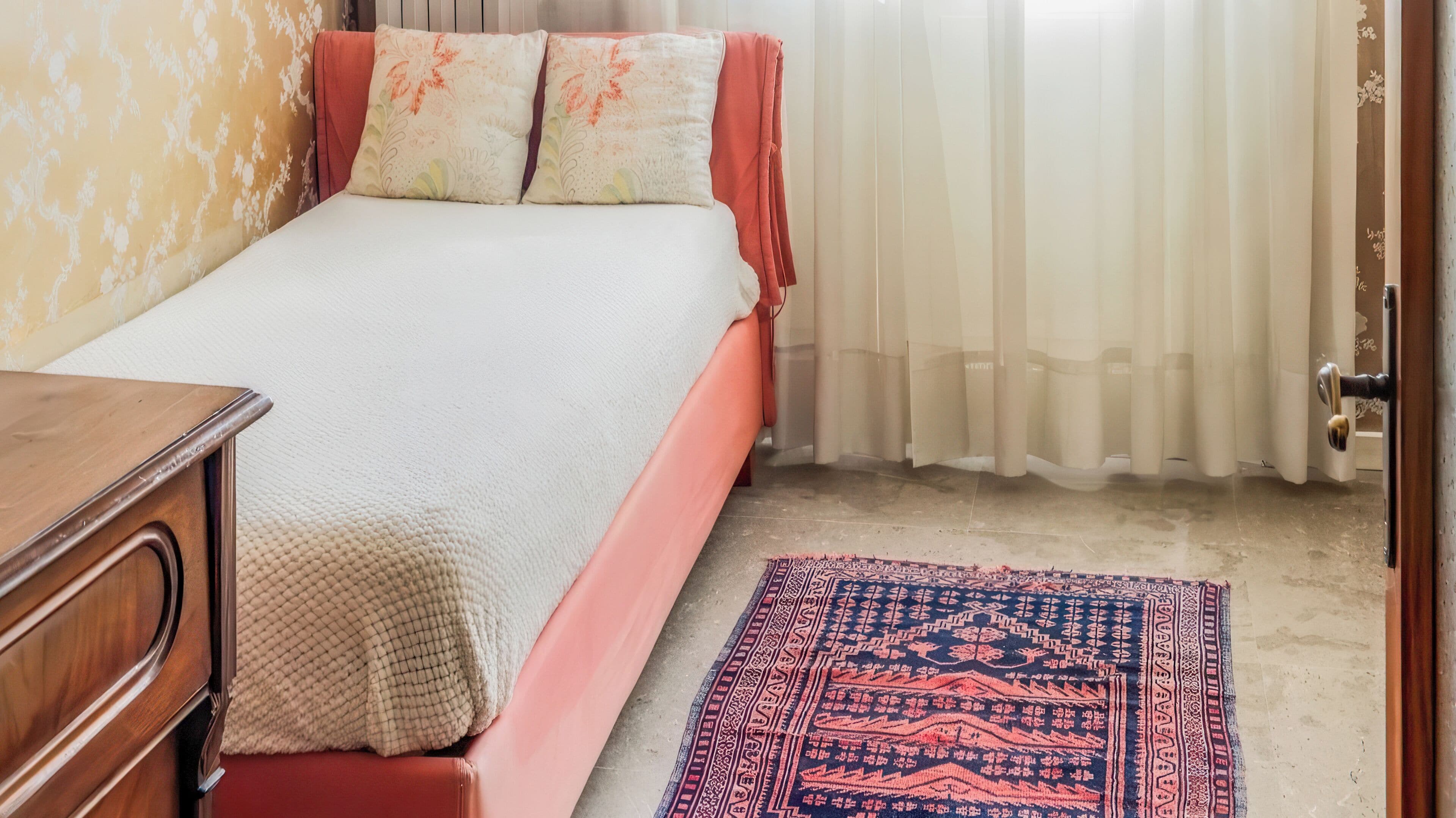 Een slaapkamer met een bed, een houten dressoir, een kamerbreed tapijt met dessin en een deur.