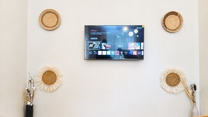 A flat-screen TV mounted on a wall with a selection of streaming services, two decorative woven wall hangings, and a vase with dried plants. - The Valley Retreat (Arusha)