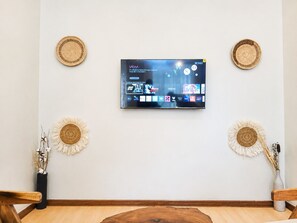 Un televisor de pantalla plana montado en la pared con acceso a servicios de streaming, dos tapices decorativos tejidos y un florero con plantas secas.