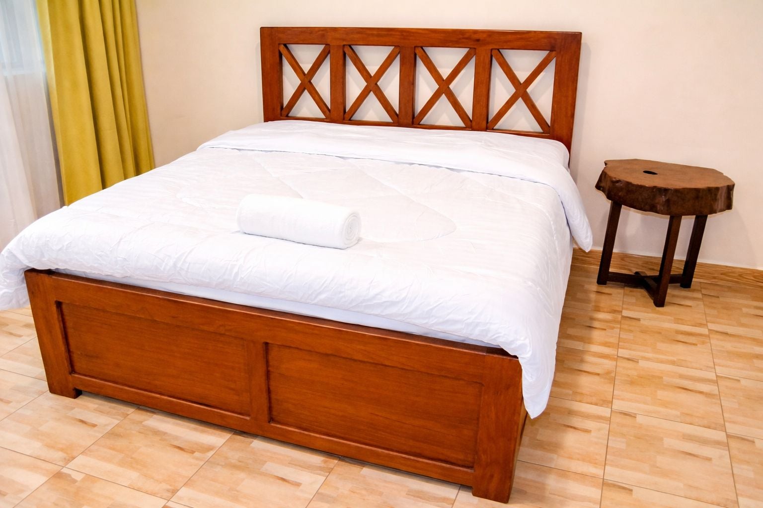A wooden bed with a cross-back design, a white bedspread, and a small round wooden stool beside it.