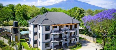 A multi-story building with a flat roof, surrounded by greenery and a mountain in the background.