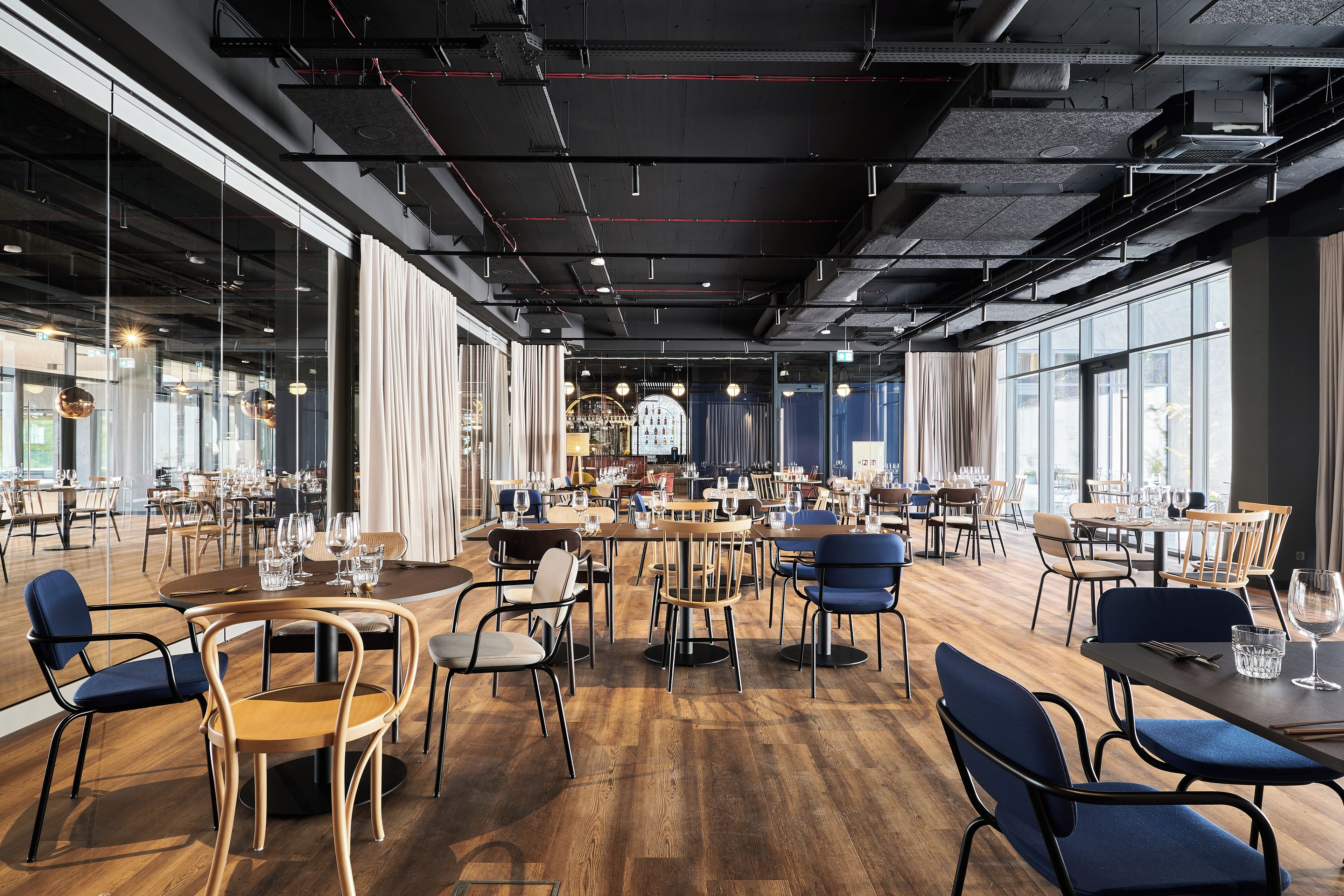 A spacious restaurant with high ceilings, exposed ductwork, and large windows. Tables are set with chairs, and the floor is wooden.