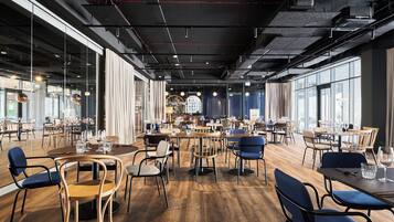A spacious restaurant with high ceilings, exposed ductwork, and large windows. Tables are set with chairs, and the floor is wooden.