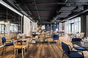 Um restaurante espaçoso com pé direito alto, dutos aparentes e grandes janelas. Mesas com cadeiras e piso de madeira.