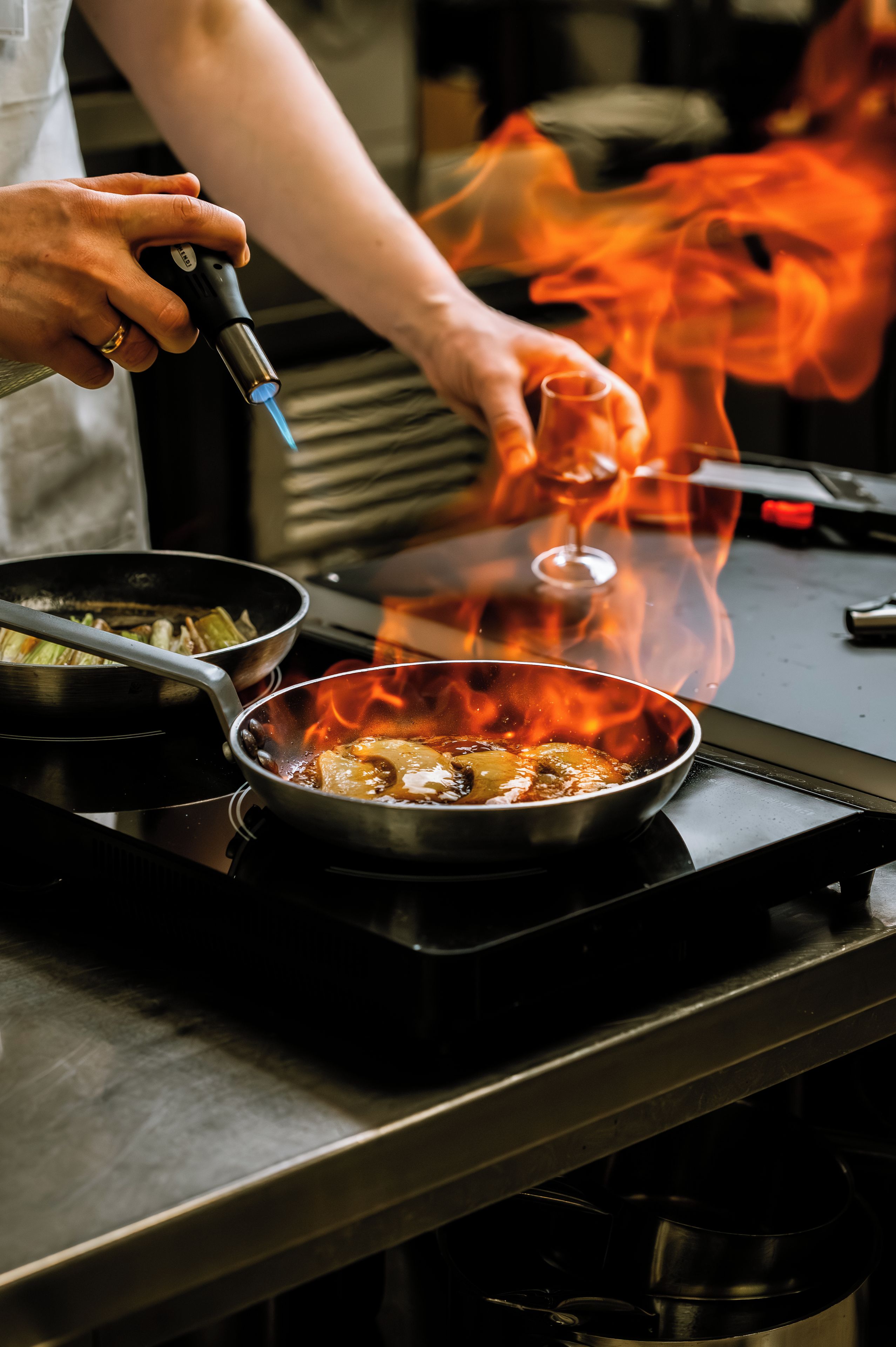 A person cooking with a pan on a stove, flames visible, and a glass nearby.