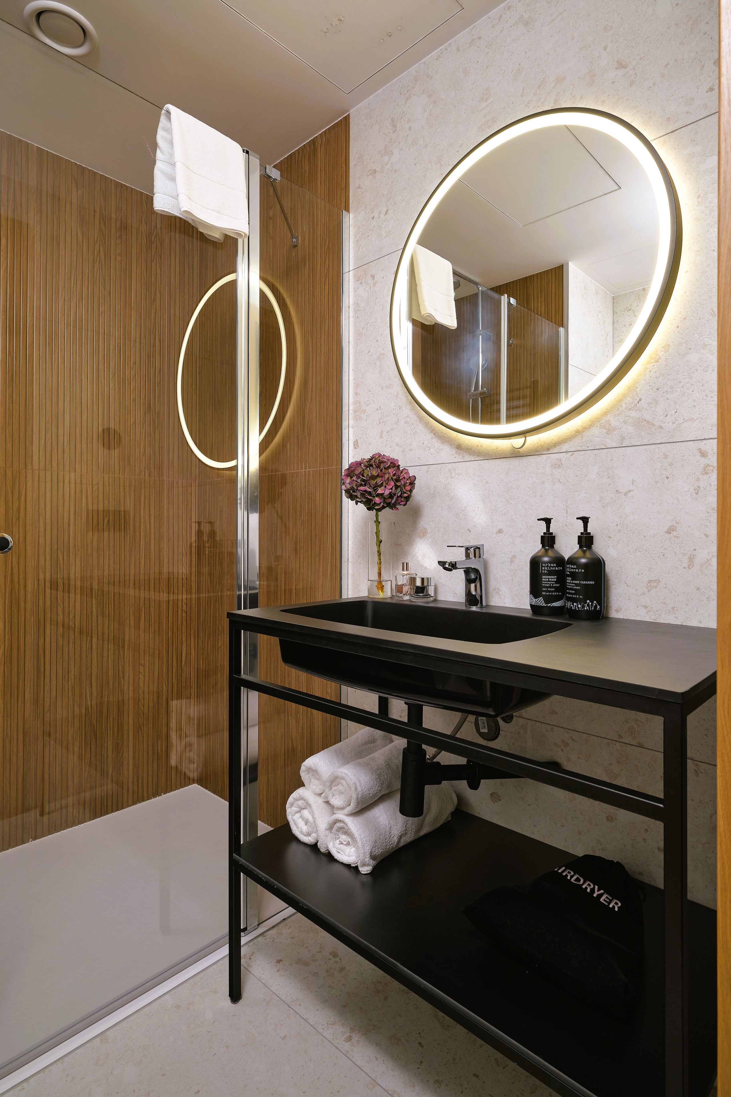 A modern bathroom with a black sink, a mirror with a circular light, a shower, and a wooden wall.