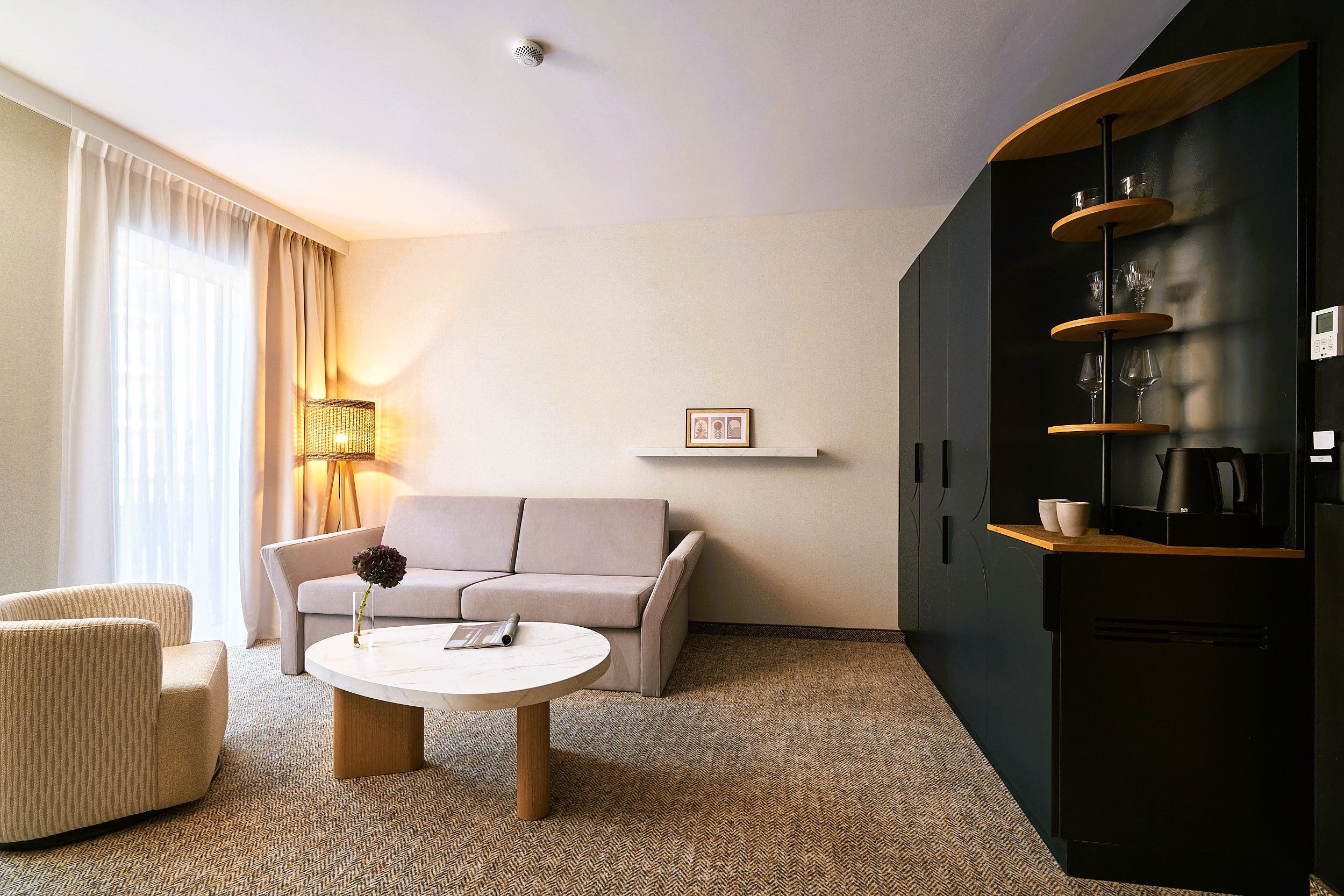 A modern hotel room with a sofa, a round coffee table, a lamp, a bookshelf, and a wall-mounted picture.