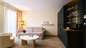 A modern hotel room with a sofa, a round coffee table, a lamp, a bookshelf, and a wall-mounted picture.