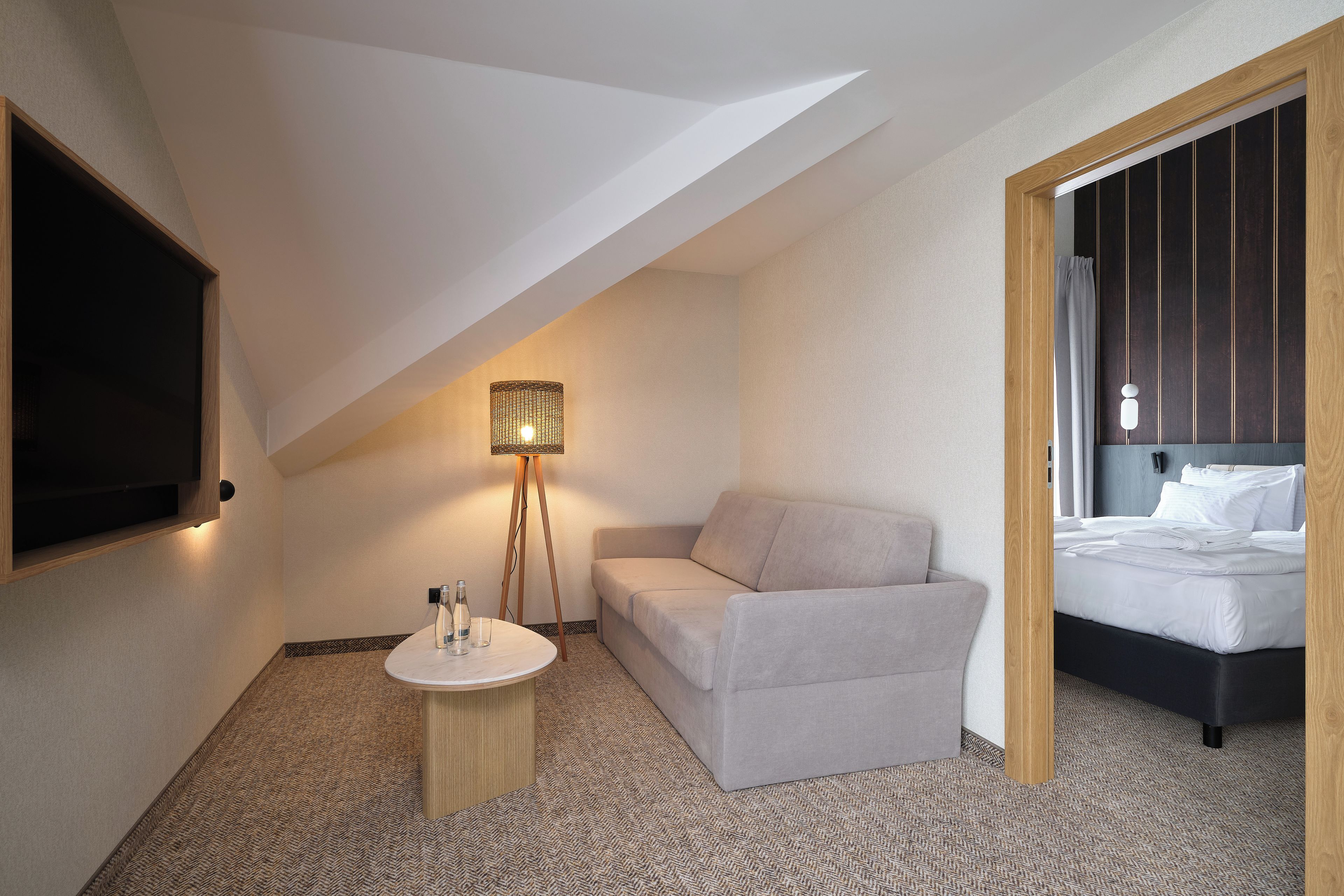 A hotel room with a sofa, a small round table, a floor lamp, a bed, and a TV mounted on the wall.