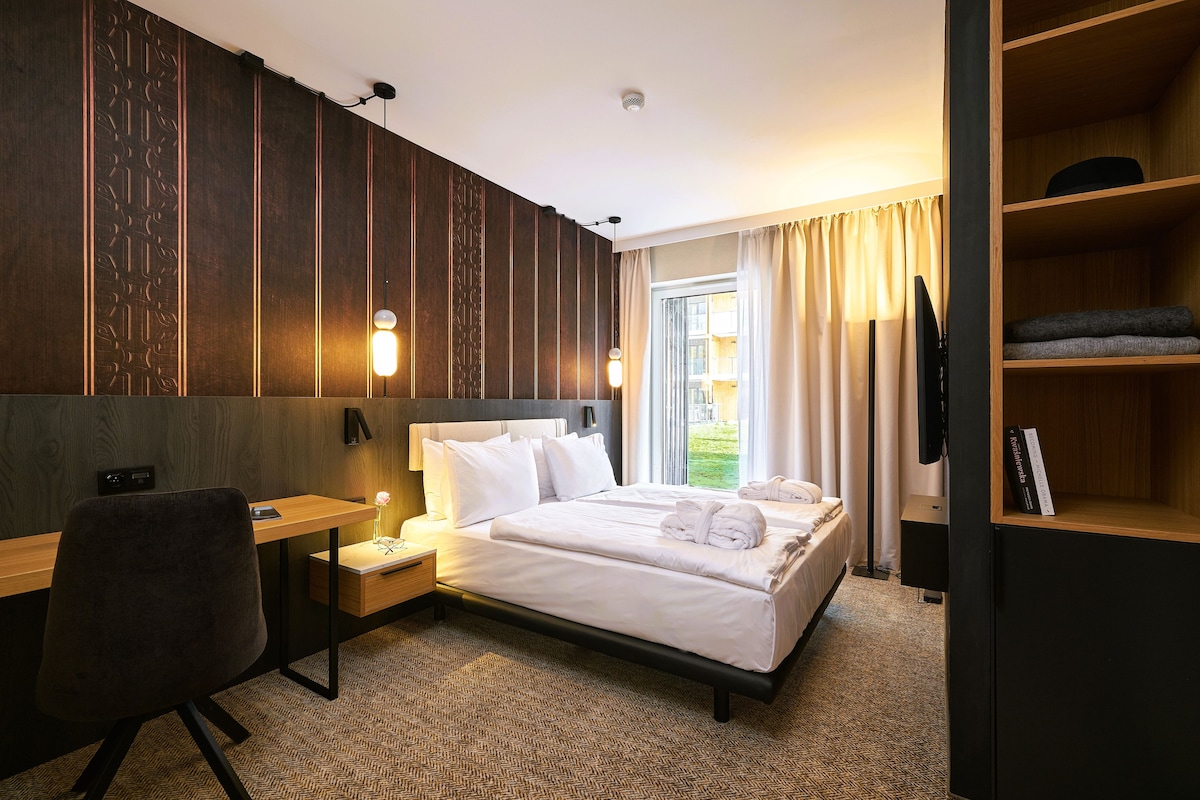 A hotel room with a bed, bedside table, desk, chair, and a bookshelf.