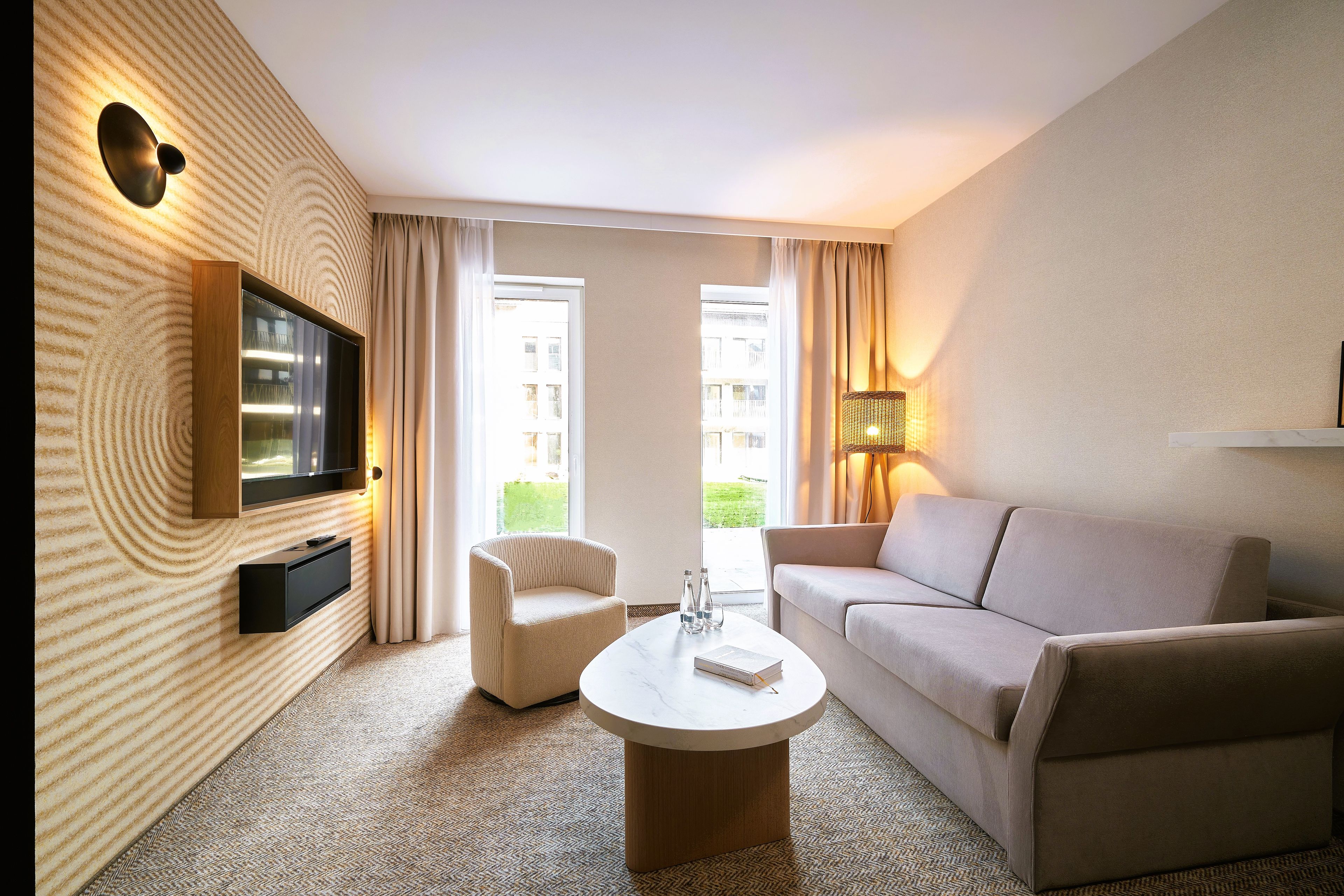 Une chambre d’hôtel équipée d’un téléviseur à écran plat fixé au mur, d’un canapé, d’un fauteuil, d’une table basse et d’une lampe.