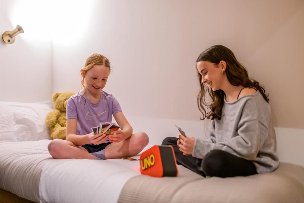 Two individuals sitting on a bed with a game board and cards, a teddy bear, and a wall-mounted light fixture.