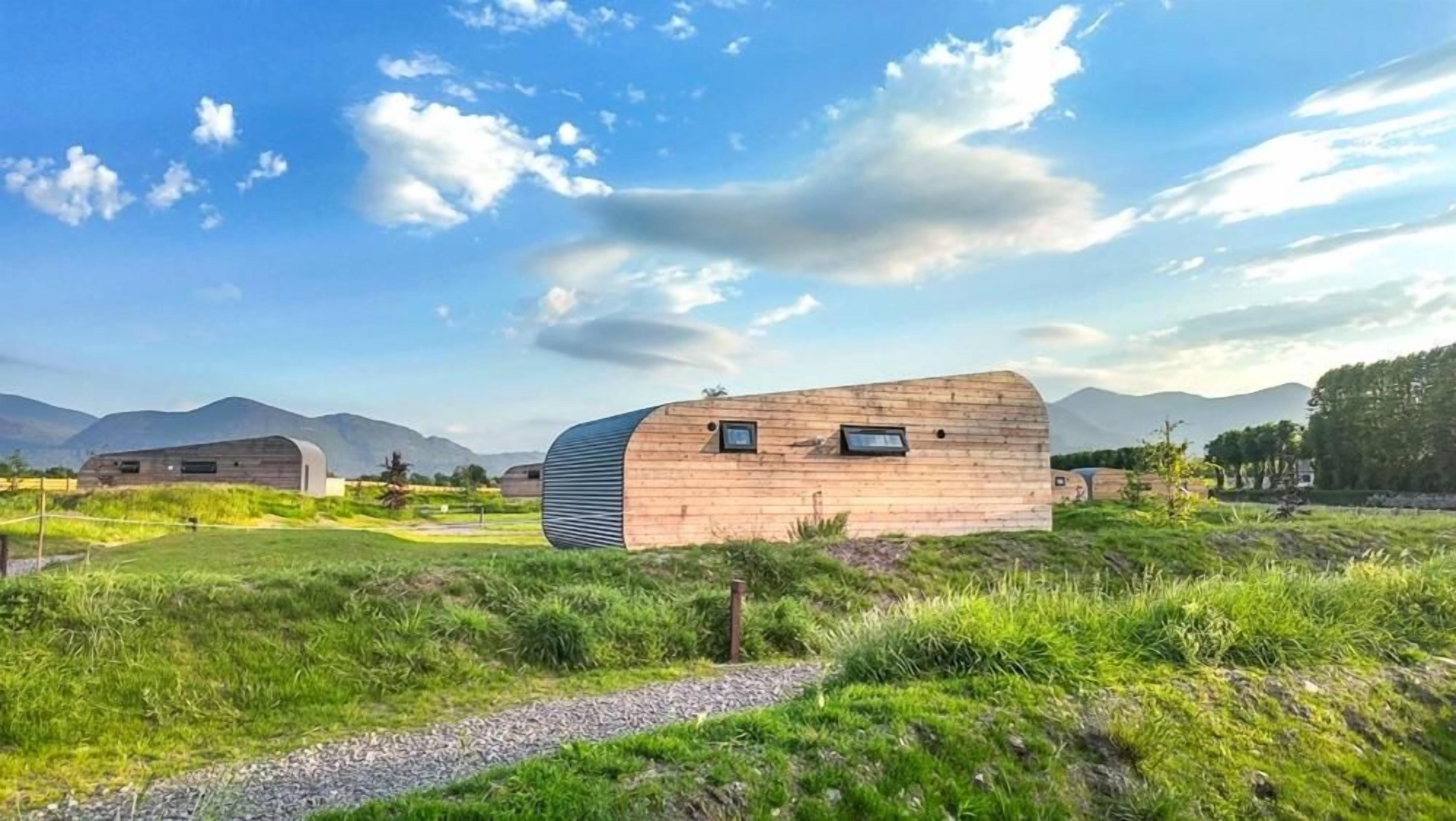 Un edificio moderno con techo curvo, rodeado por un paisaje cubierto de césped y montañas al fondo.