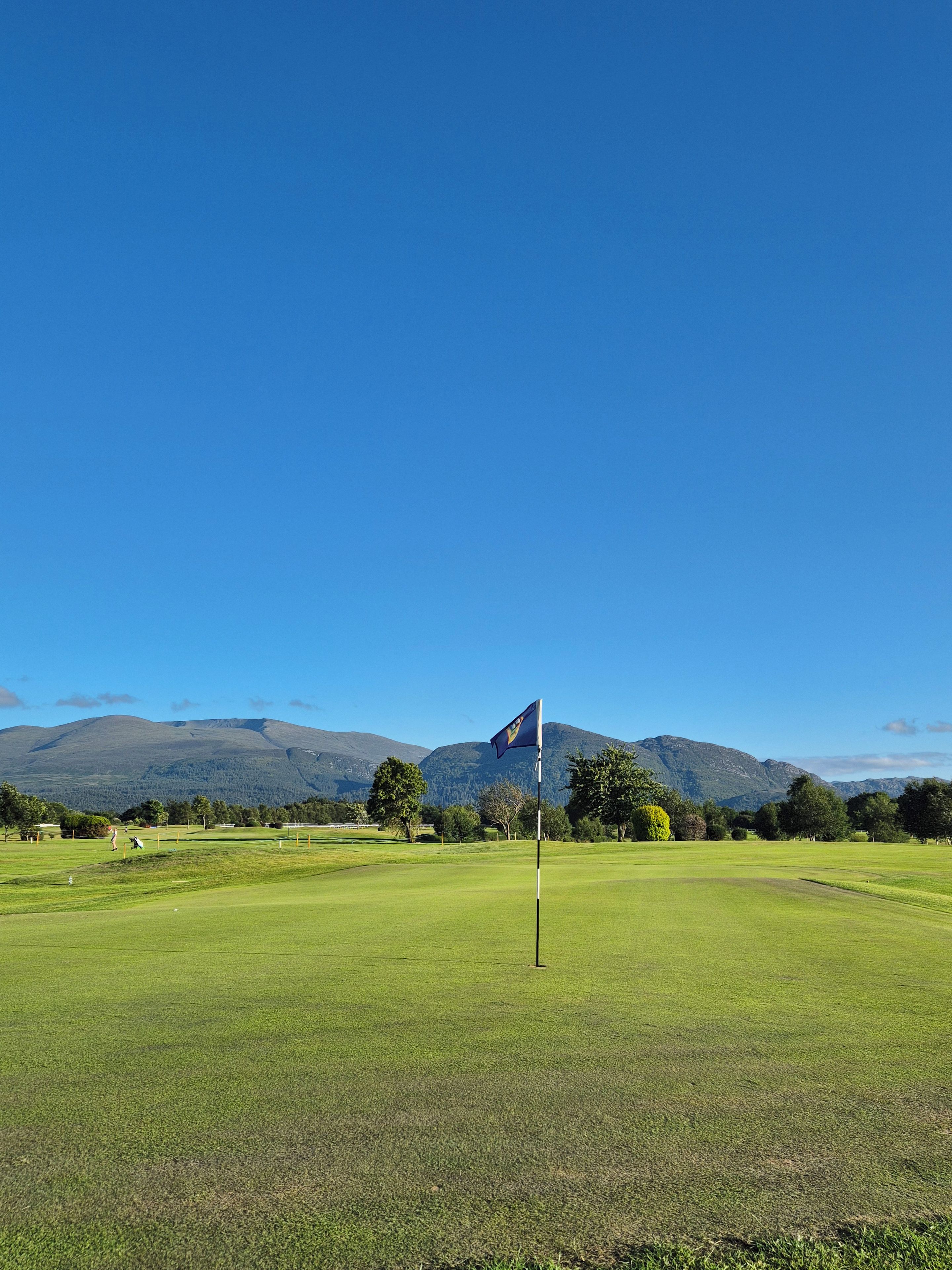 Un campo de golf con una bandera en un asta, rodeado de hierba verde y árboles, con montañas al fondo.