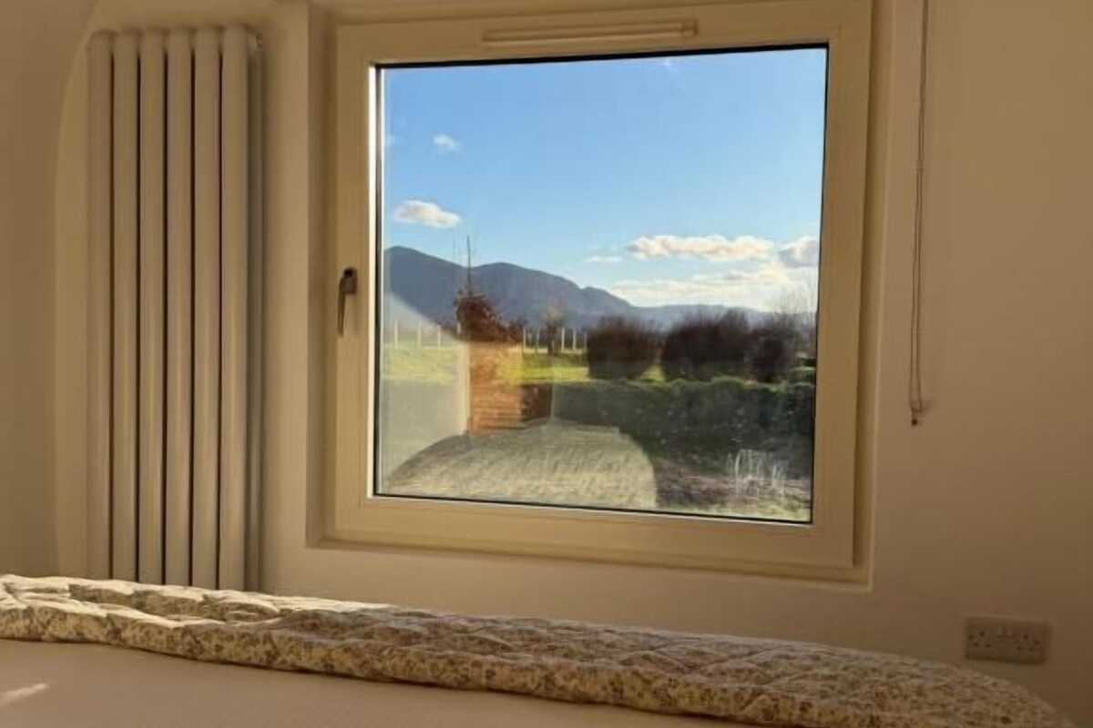 Ein Schlafzimmer mit großem Fenster, einem Holzbalken an der Decke und einem Bett mit gemustertem Kopfteil.