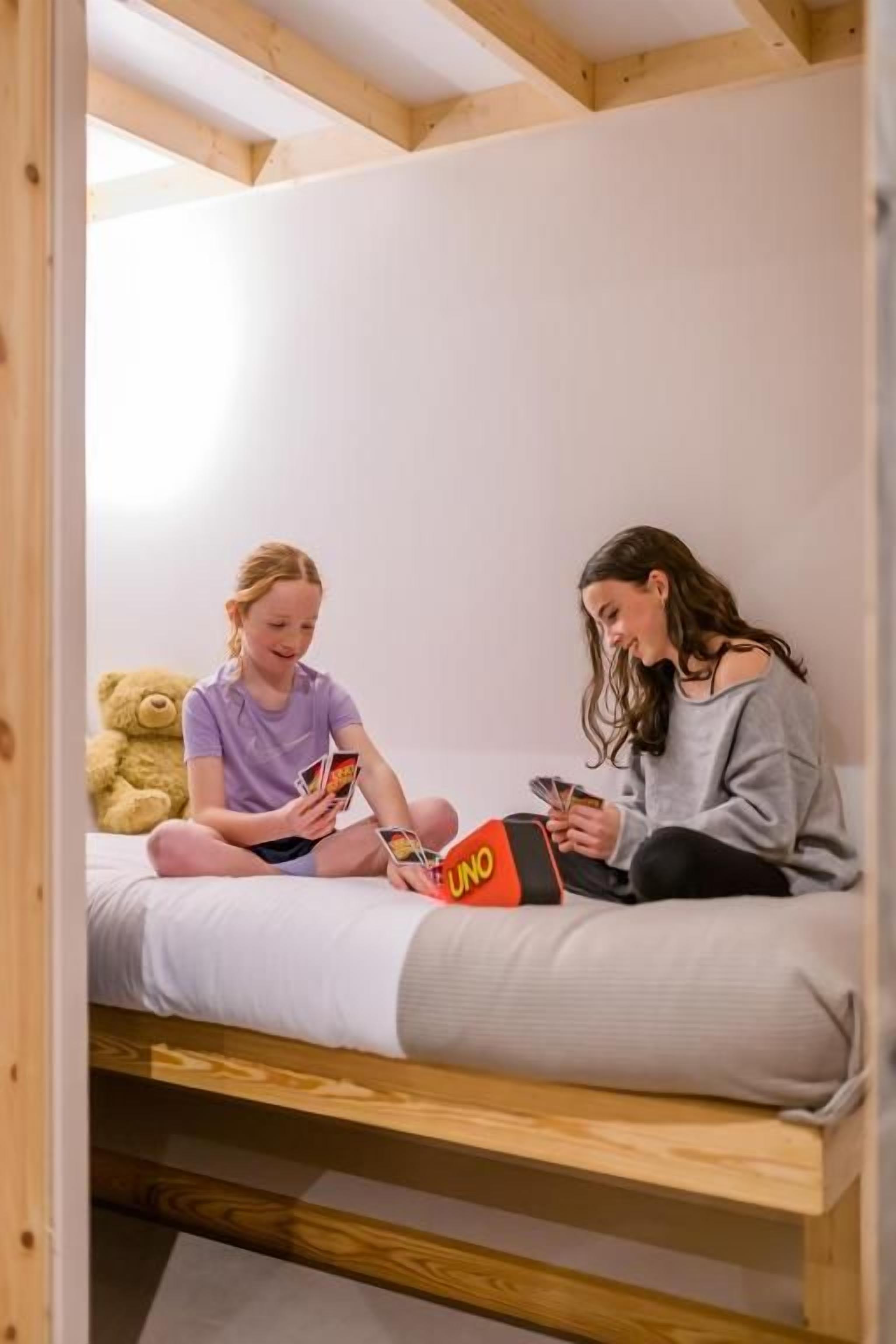 Dos personas sentadas en una cama litera, con un osito de peluche y una caja del juego UNO.