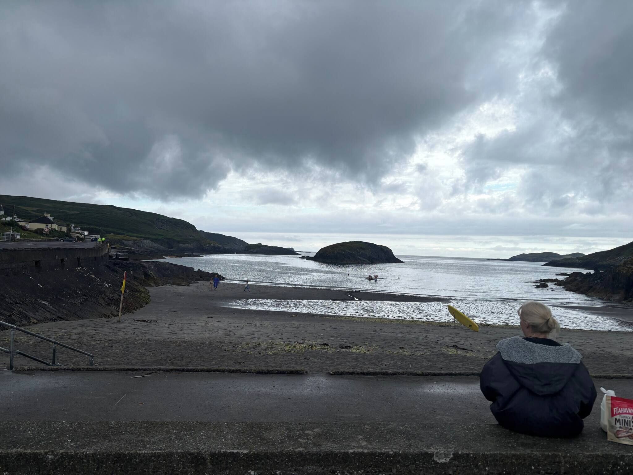 A person sitting on a concrete ledge overlooking a beach with a rocky coastline and overcast sky.
