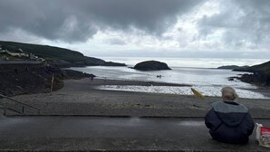A person sitting on a concrete ledge overlooking a beach with a rocky coastline and overcast sky. - Charming 4-bedroom House in Skibbereen - Pet Friendly Retreat (Skibbereen)