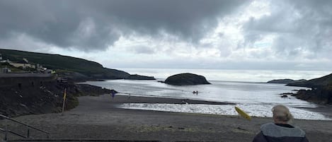 Eine Person sitzt auf einer Betonstufe und blickt auf einen Strand mit felsiger Küste unter einem bewölkten Himmel.