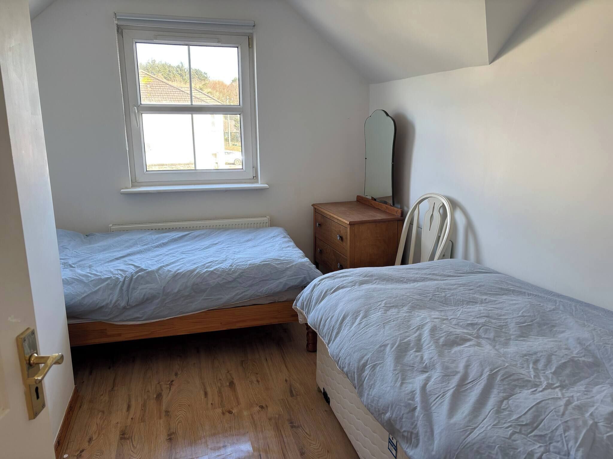 A bedroom with two single beds, a wooden dresser, a mirror, and a window with a view of the outside.