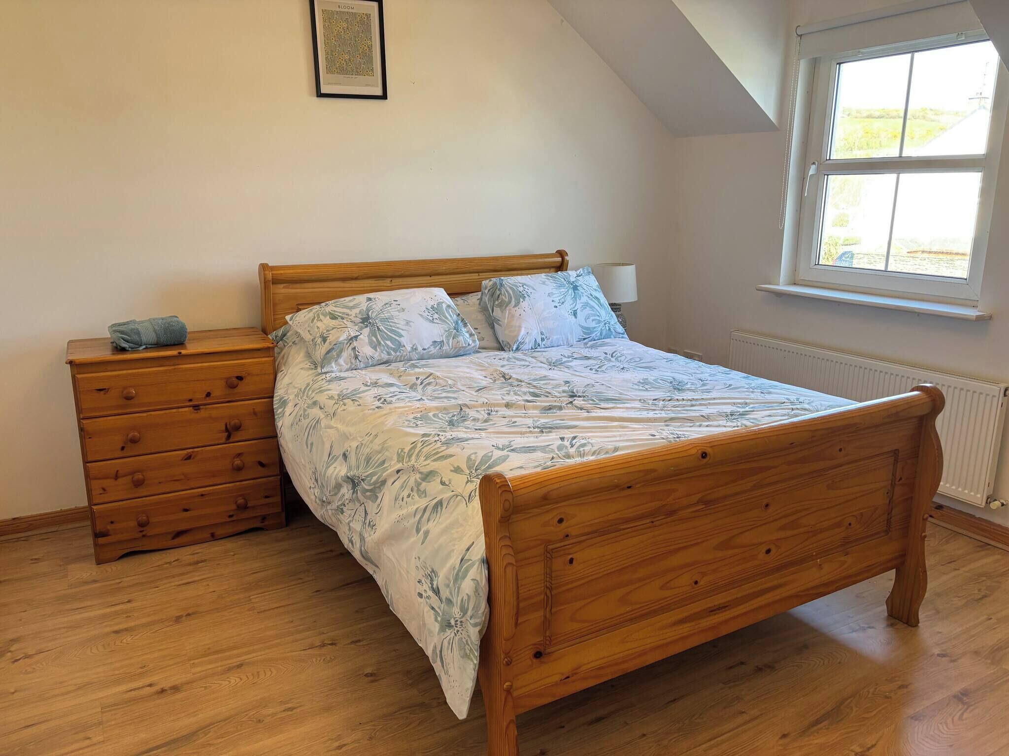 A bedroom with a wooden bed, a wooden dresser, a framed picture on the wall, and a window with a view.