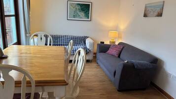 A cozy living room with a wooden dining table, white chairs, a grey sofa, and a small lamp.