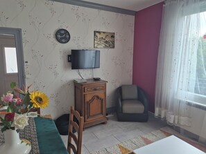 A living room with a clock, a flat-screen TV, a sofa, a chair, and a table with a vase of flowers. - Large village house, 90 m² – Sleeps 10 – Lac du Der – 8 km from Saint-Dizier (Hallignicourt)