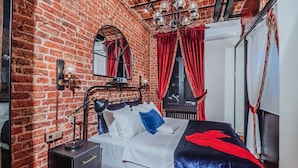A room with a brick wall, a bed with white bedding, a black metal headboard, a black nightstand with a lamp, and red curtains. - Allegory of Cihangir 2B (İstanbul)