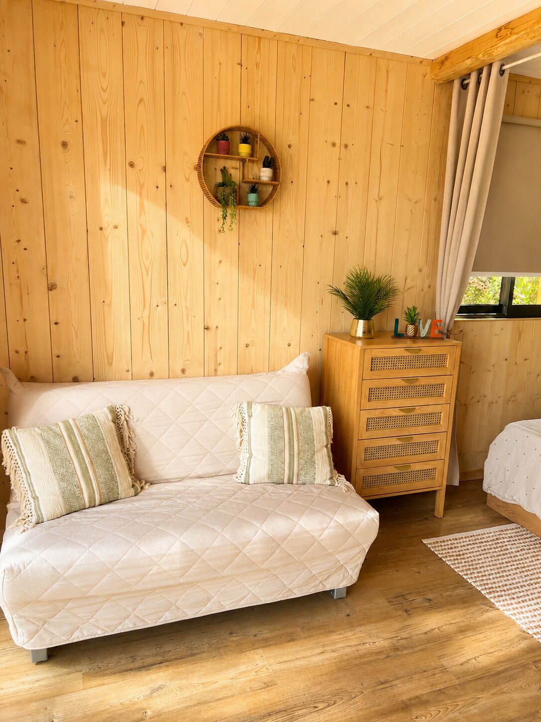 Une pièce chaleureuse avec un mur en bois, une étagère ronde ornée de plantes, une commode en bois et un lit recouvert d’une courtepointe blanche.