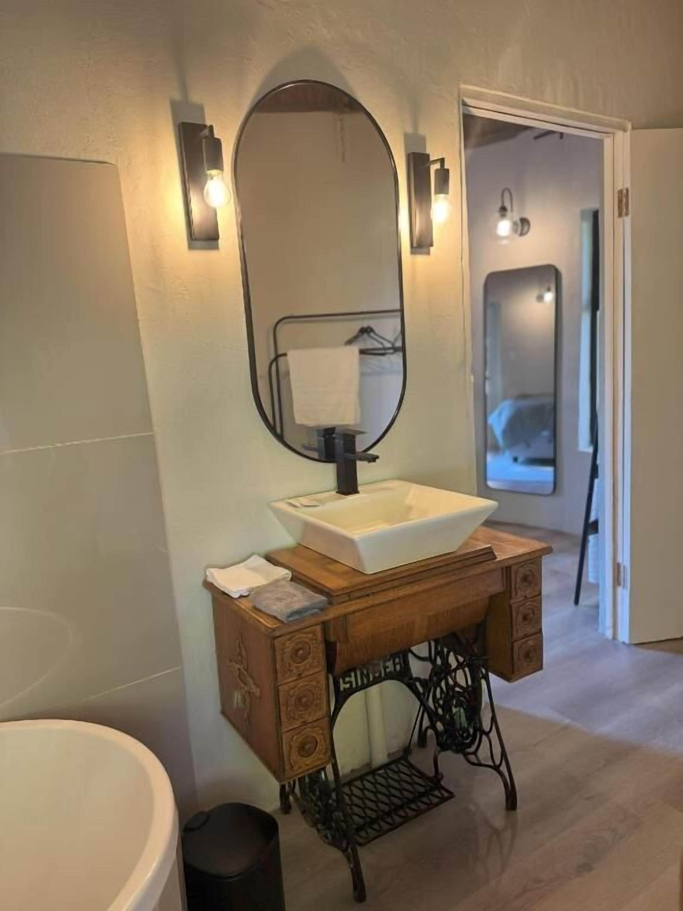 A bathroom with a white sink, a wooden vanity, a mirror, and a black sewing machine.