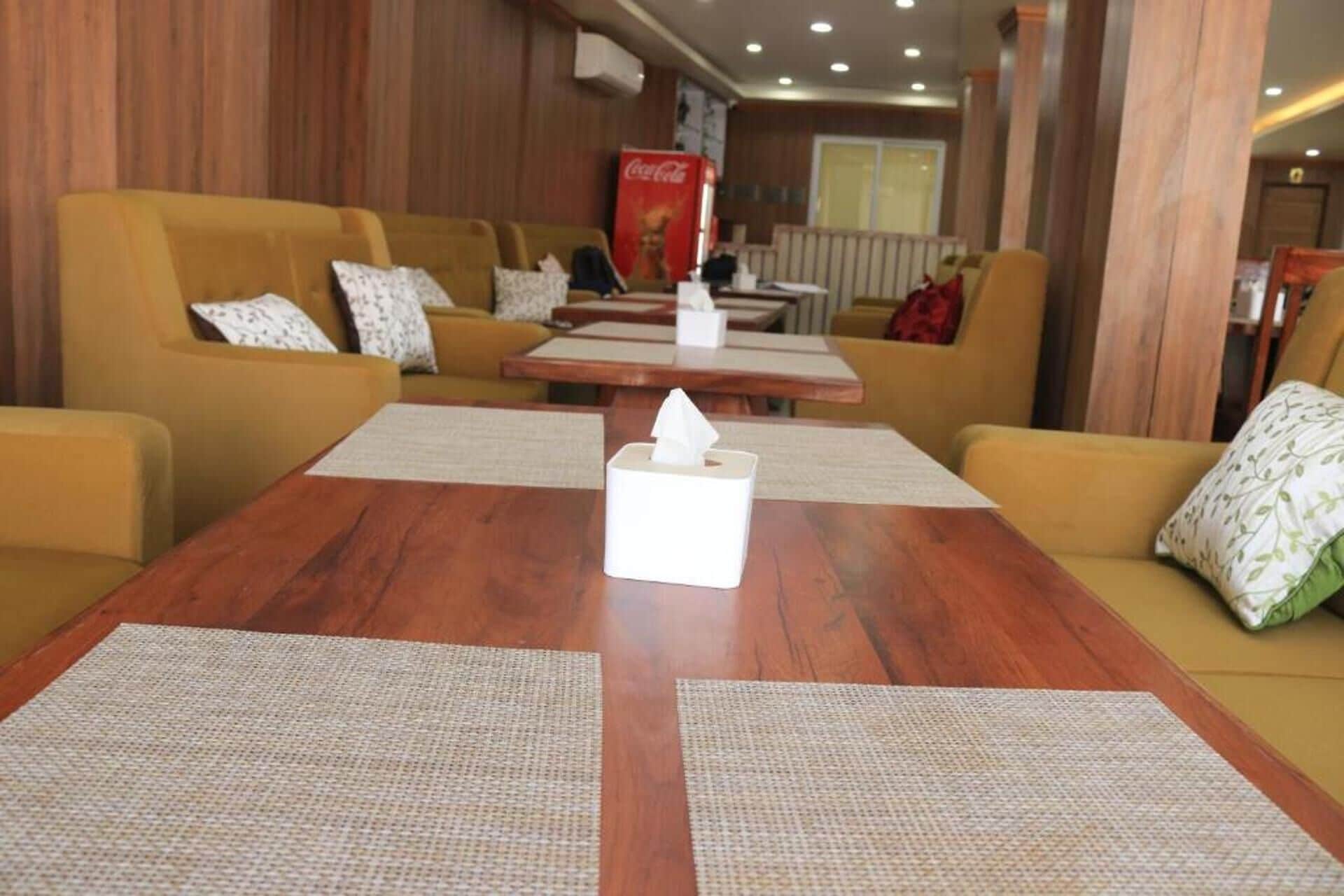 Eine Hotellobby mit Holzwandverkleidung, gelben Sofas mit gemusterten Kissen, einem Holztisch mit Taschentuchbox und einem Coca-Cola-Kühlschrank.