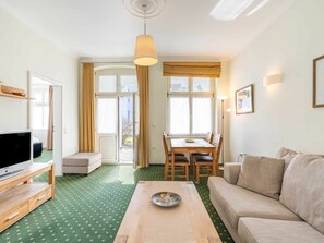 A room with a television on a stand, a sofa, a dining table with chairs, and a large window with curtains. - Holiday Apartment With 1 Bedroom (Heringsdorf)