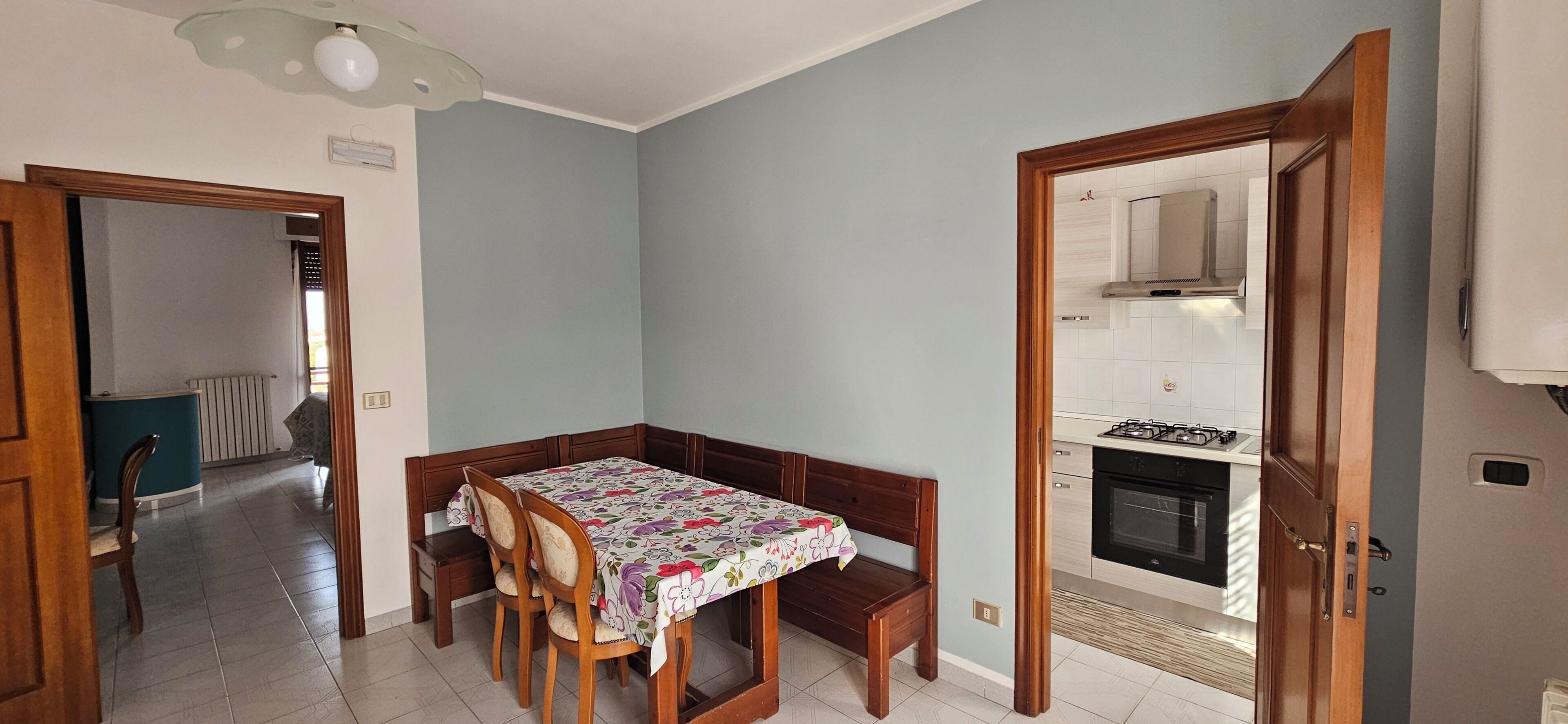Un coin repas avec une table et des chaises, une cuisine équipée d’une cuisinière et d’un four, et un couloir menant à une autre pièce.