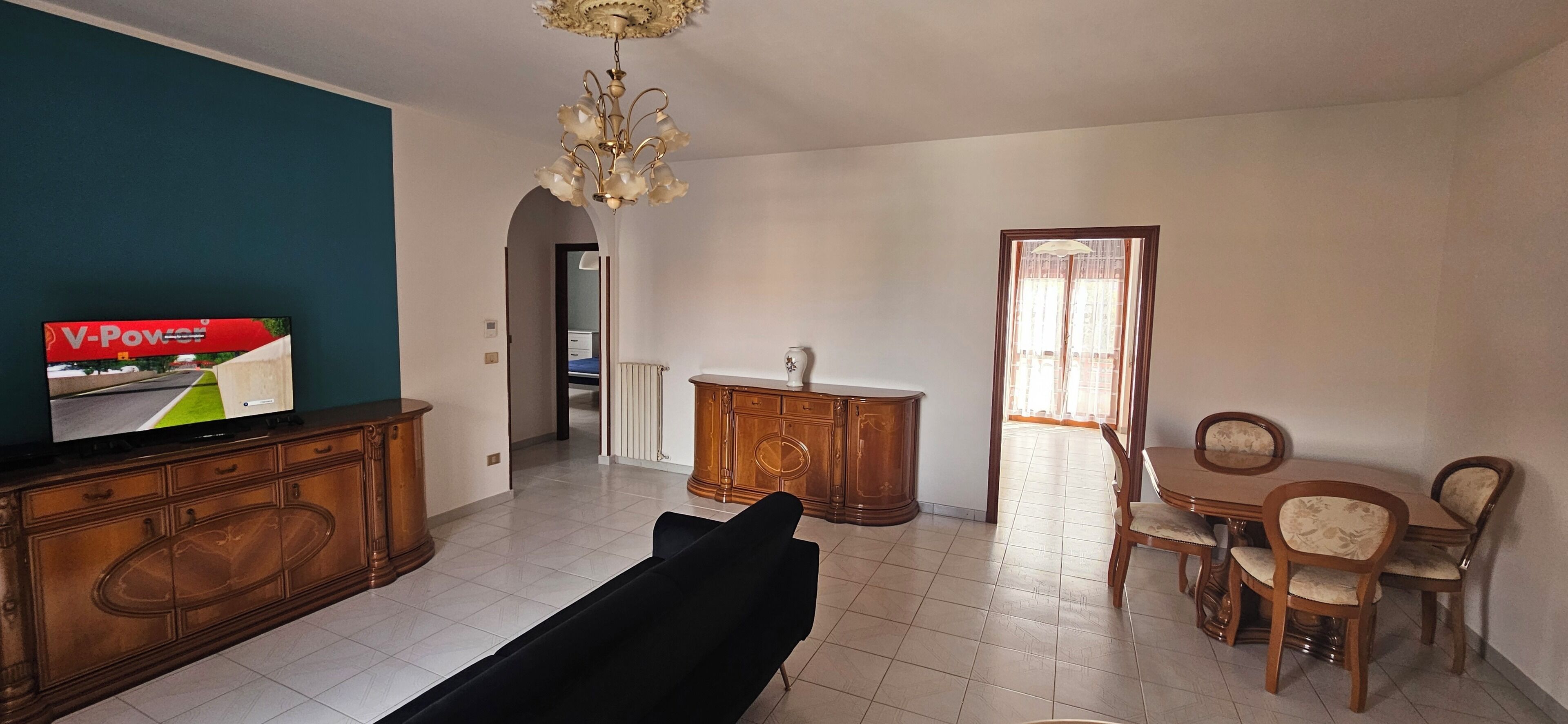 Un salon comprenant un téléviseur, un canapé, une table à manger avec des chaises, un lustre et un buffet.