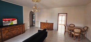 A living room with a television, a sofa, a dining table with chairs, a chandelier, and a cabinet.