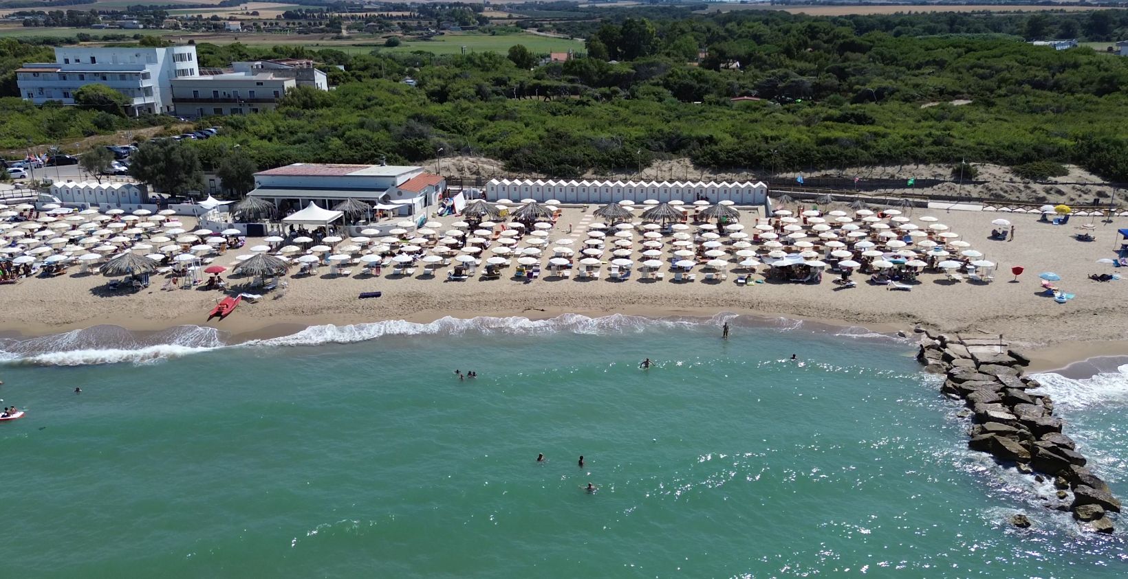 A beach with numerous umbrellas and sun loungers, a rocky breakwater, and a building with a red roof in the background.